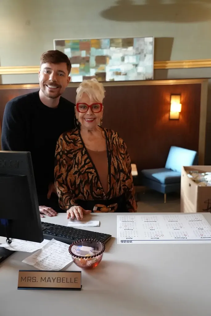 Jimmy Donaldson poses with a woman in a patterned jacket and red-framed glasses behind a white desk. The desk has a computer, scattered papers, a 2026 calendar, a candy bowl, and a nameplate that reads "Mrs. Maybelle." A small sitting area is visible in the background.