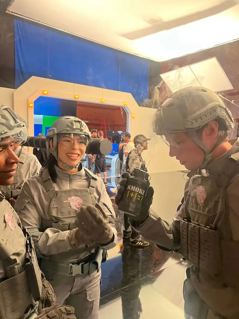Three people on a film set wearing gray tactical vests and helmets inspect a prop explosive on a film set. A woman in the foreground smiles at the camera. Film crew and lighting equipment are visible in the background.