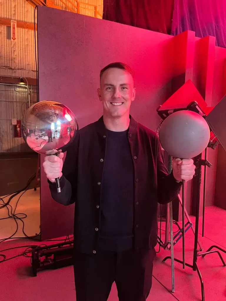 A smiling man in black clothing stands on a film set holding two production lights—a chrome reflective sphere light in his left hand and a frosted globe light with a stand in his right hand. The set is bathed in pink and red lighting. Cables and equipment are visible on the floor.